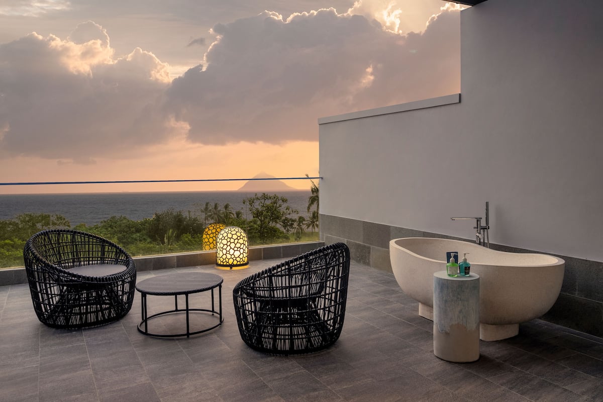 Un espacio exterior moderno con dos sillones de mimbre y una mesa, una bañera grande, y vistas al océano y las nubes.