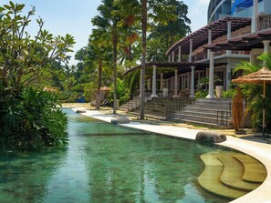 Une piscine aux eaux cristallines, avec des marches menant à un espace abrité, le tout entouré d’une végétation luxuriante et de palmiers.