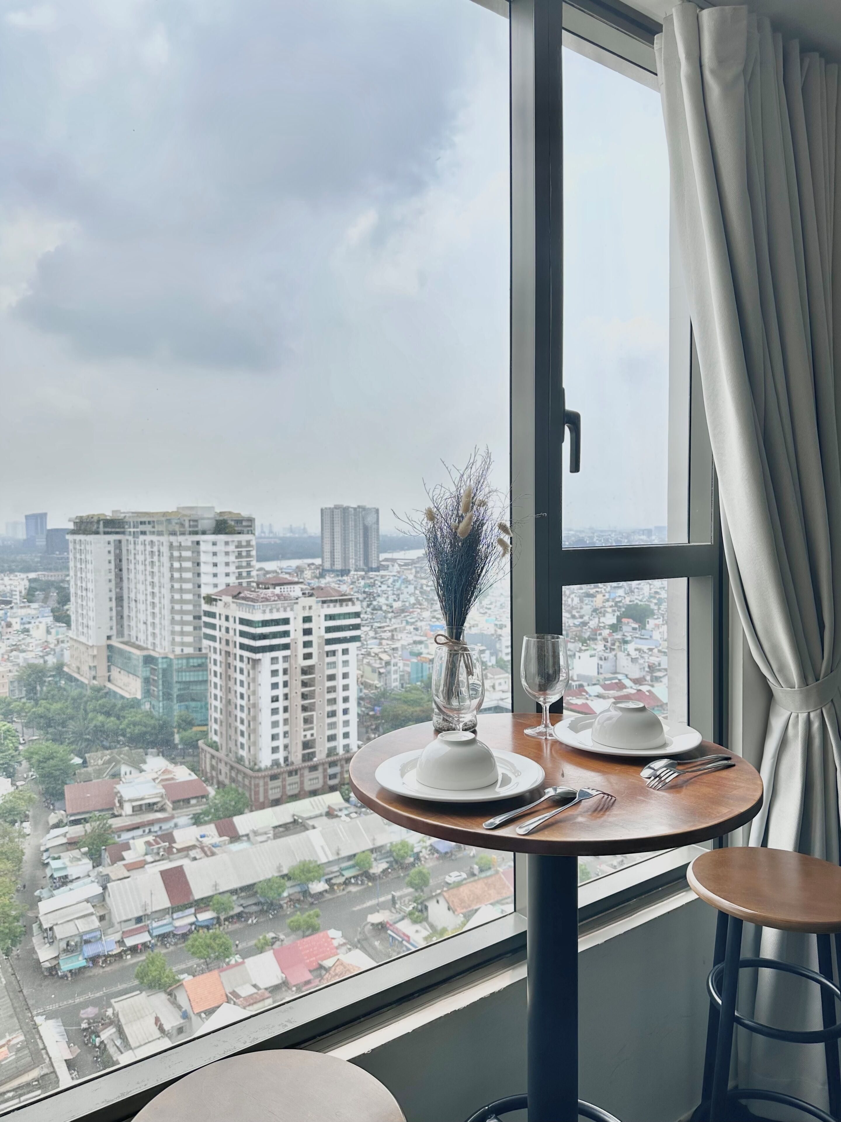 Habitación en un rascacielos con vista a la ciudad, una mesa redonda con un florero y una ventana con cortinas.