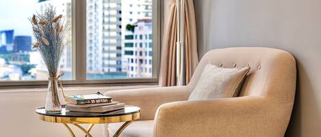 A modern living room with a view of a cityscape, a beige armchair, a small round table with books and a vase, and a yellow ottoman.