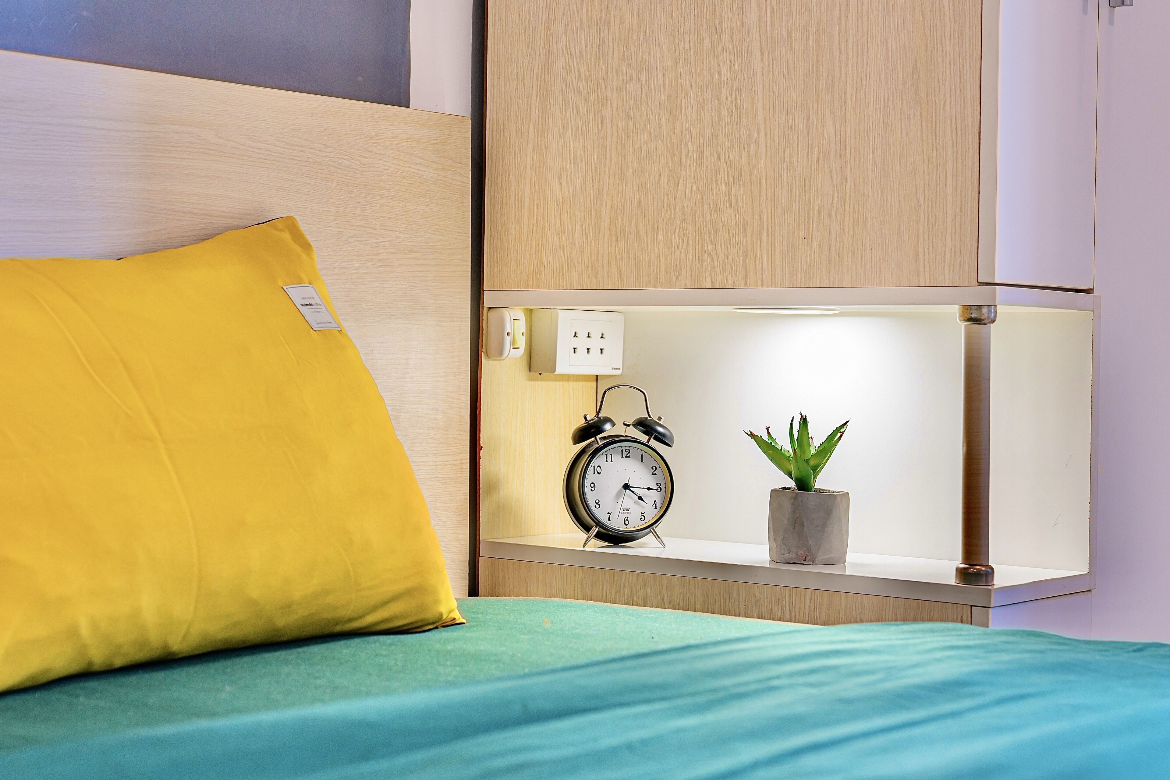 Un lit avec un oreiller jaune, une couvre-lit verte, une table de chevet avec une petite plante en pot et un réveil fixé au mur.