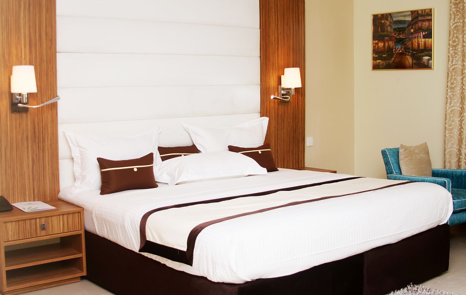 A hotel room with a large bed, two bedside tables, a nightstand, a blue armchair, and a painting on the wall.