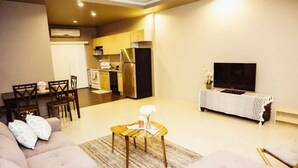 A modern living room with a sofa, a coffee table, and a TV. There is a kitchen area with a refrigerator and a dining table with chairs. - Tumon Belair (Tamuning)