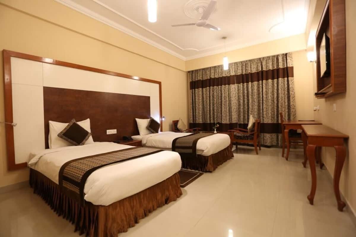 Quarto de hotel com duas camas, uma escrivaninha, uma cadeira, ventoinha no teto e televisão.