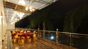 Une salle de banquet où les tables sont dressées pour un événement formel, avec un sol en marbre poli et une balustrade en verre donnant sur un palmier.
