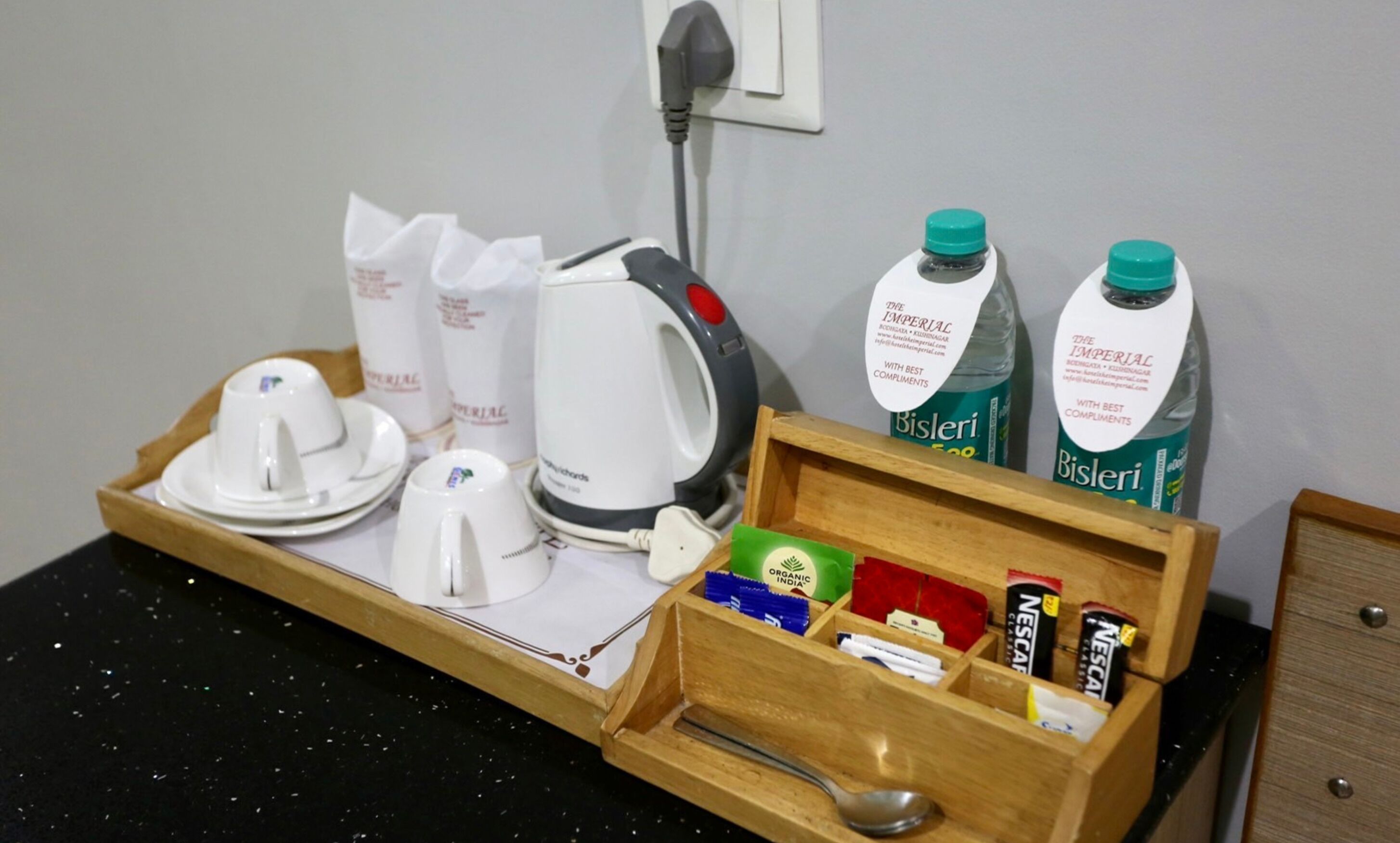 Habitación de hotel con una bandeja de madera que contiene una tetera, tazas y botellas de agua Bisleri.
