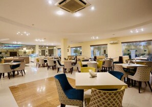A spacious dining area with multiple tables and chairs, a central air conditioning unit, and large windows providing natural light. - The Imperial Bodhgaya (Gaya)