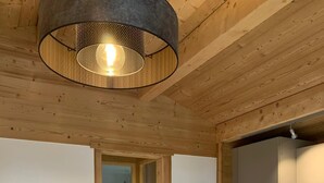Une pièce chaleureuse avec un plafond en bois, un lit recouvert d’une housse à motifs, une petite table avec une lampe et un coin cuisine avec des placards.