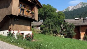 Un chalet en bois doté d’un balcon, entouré d’une végétation luxuriante et avec des montagnes en toile de fond.