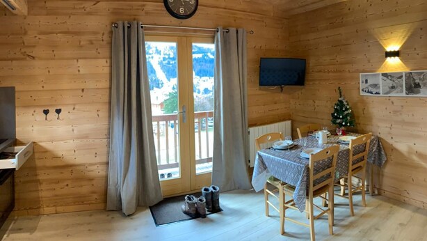 L'intérieur d'un chalet en bois, avec une table à manger dressée pour deux personnes, un téléviseur fixé au mur et une horloge également accrochée au mur.