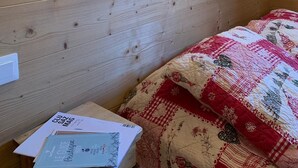 Une pièce aux murs lambrissés, avec deux lampes fixées au mur, un lit recouvert d’une courtepointe et une table de chevet sur laquelle se trouvent des papiers et un livre.