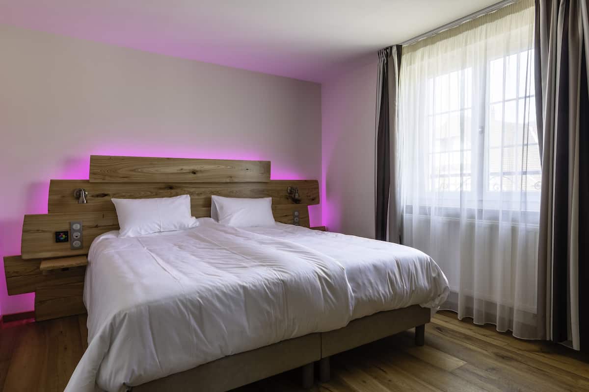 A neatly made bed with white linens in a room with wooden flooring, a wooden headboard, and a window with sheer curtains.