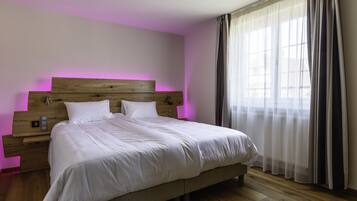 A neatly made bed with white linens in a room with wooden flooring, a wooden headboard, and a window with sheer curtains.