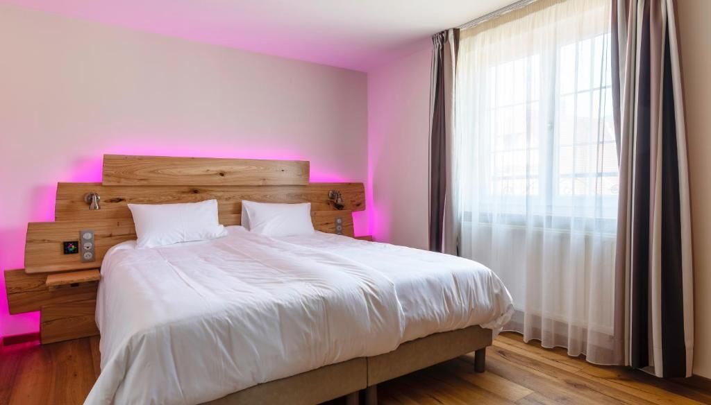 Een moderne hotelkamer met een houten hoofdeinde, een groot bed, een nachtkastje en een raam met dunne gordijnen.