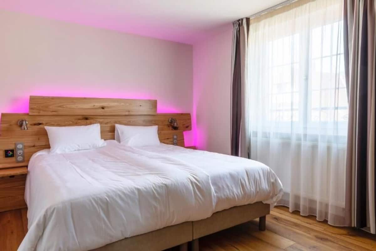 A modern hotel room with a wooden headboard, a large bed, a bedside table, and a window with sheer curtains.