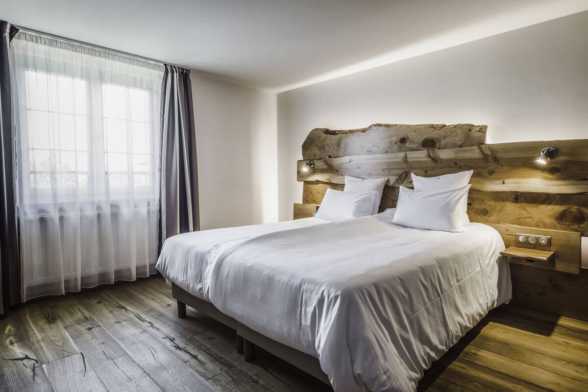 A bedroom with a wooden headboard, a large bed with white linens, a bedside table with a lamp, and a window with sheer curtains.