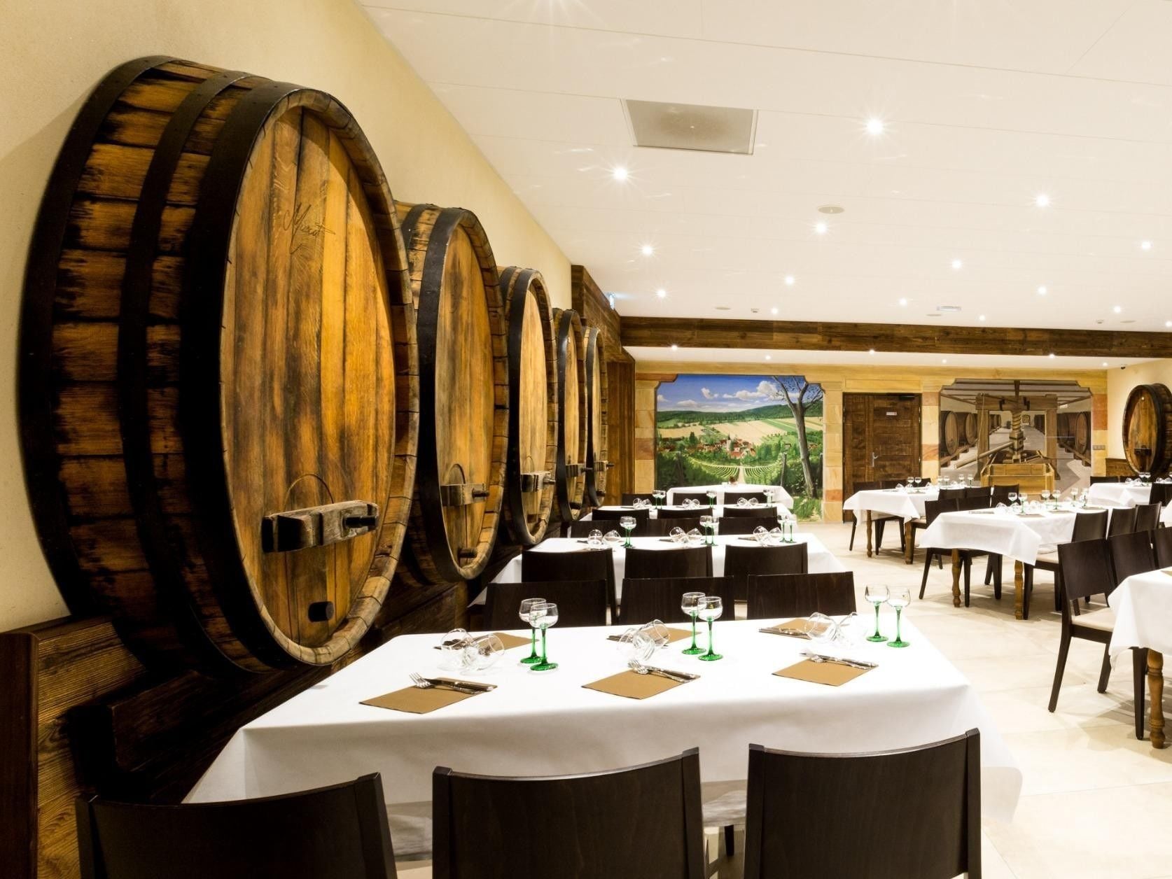 A restaurant with multiple tables, wooden barrels on the wall, and a scenic mural in the background.