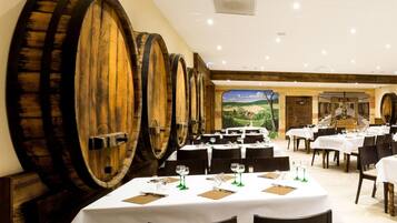 A restaurant with multiple tables, wooden barrels on the wall, and a scenic mural in the background.