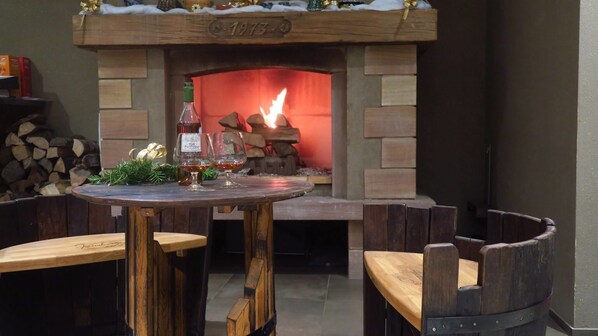 An outdoor seating area with a rustic wooden table and benches, a fireplace with a fire, and a stack of firewood in the background. - Logis Hotel Restaurant Keimberg (Cleebourg)