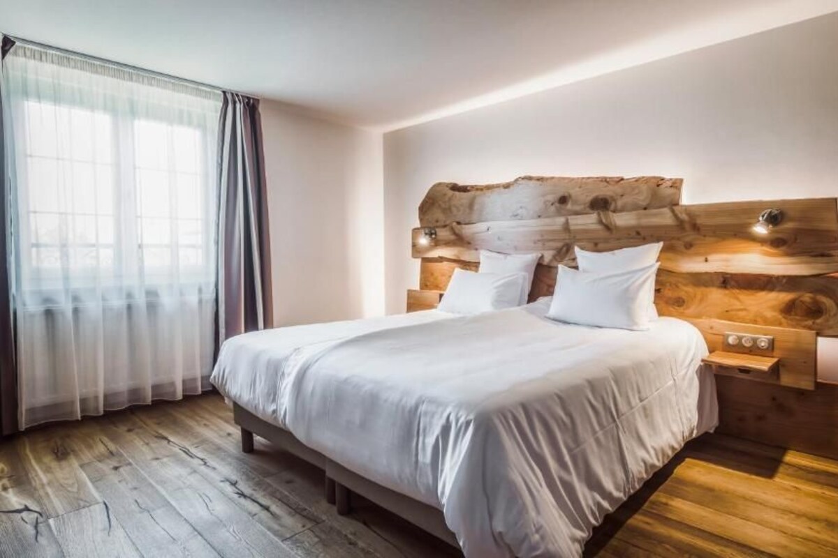 A bedroom with a wooden headboard, a bed with white bedding, wooden nightstands, and a large window with sheer curtains.