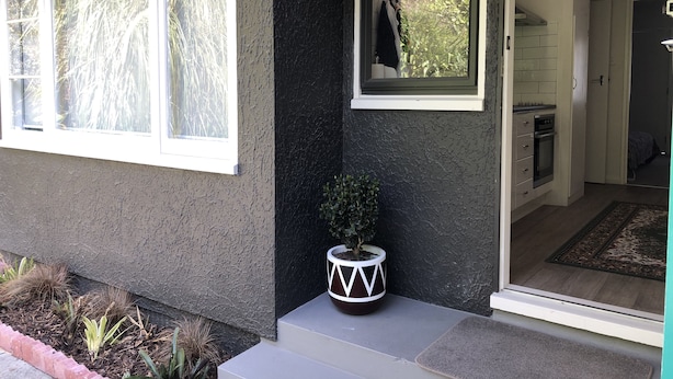 A house exterior with a dark gray stucco wall, white trim windows, a small potted plant, and a visible kitchen area with a stove and cabinets.
