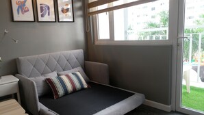 Ein modernes Schlafzimmer mit einer grauen Schlafcouch, einem kleinen Nachttisch und einem Fenster mit Blick auf einen Spielplatz.