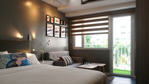 Ein modernes Hotelzimmer mit einem großen Bett, einem Schlafsofa, einem kleinen Tisch und einem Balkon mit Blick auf Grünflächen.