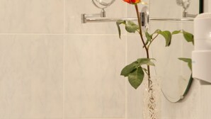 Une salle de bain avec un mur en marbre blanc, un miroir rond et un vase contenant une seule rose orange.