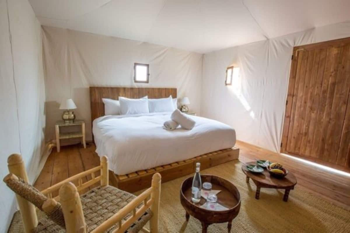 A bedroom with a wooden bed, a small bedside table, a chair, a round table with a bottle and glasses, and a wooden wardrobe.