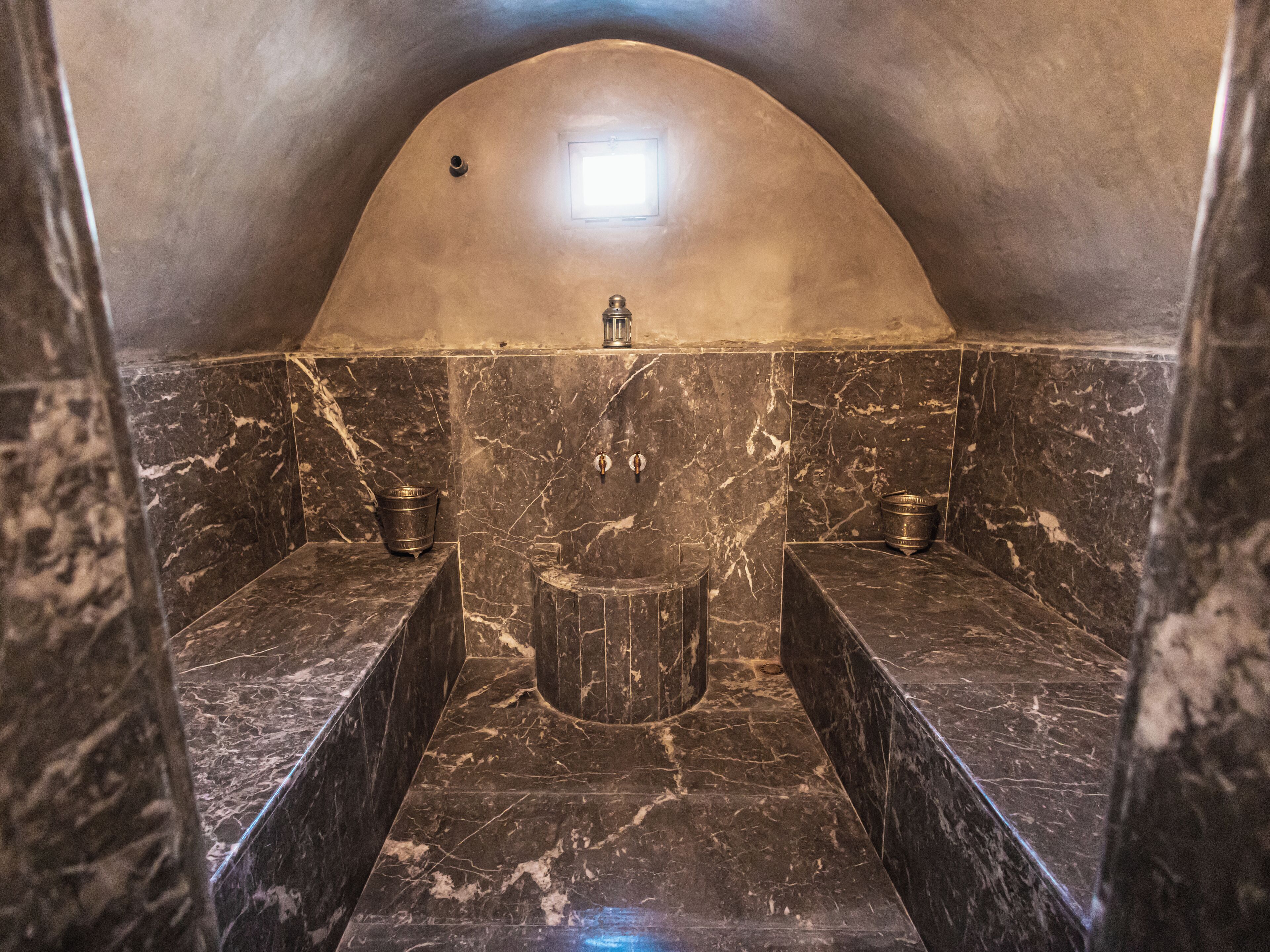 Une salle de bain aux murs en pierre, avec un lavabo central et deux lavabos latéraux, chacun doté d’un receveur en métal.