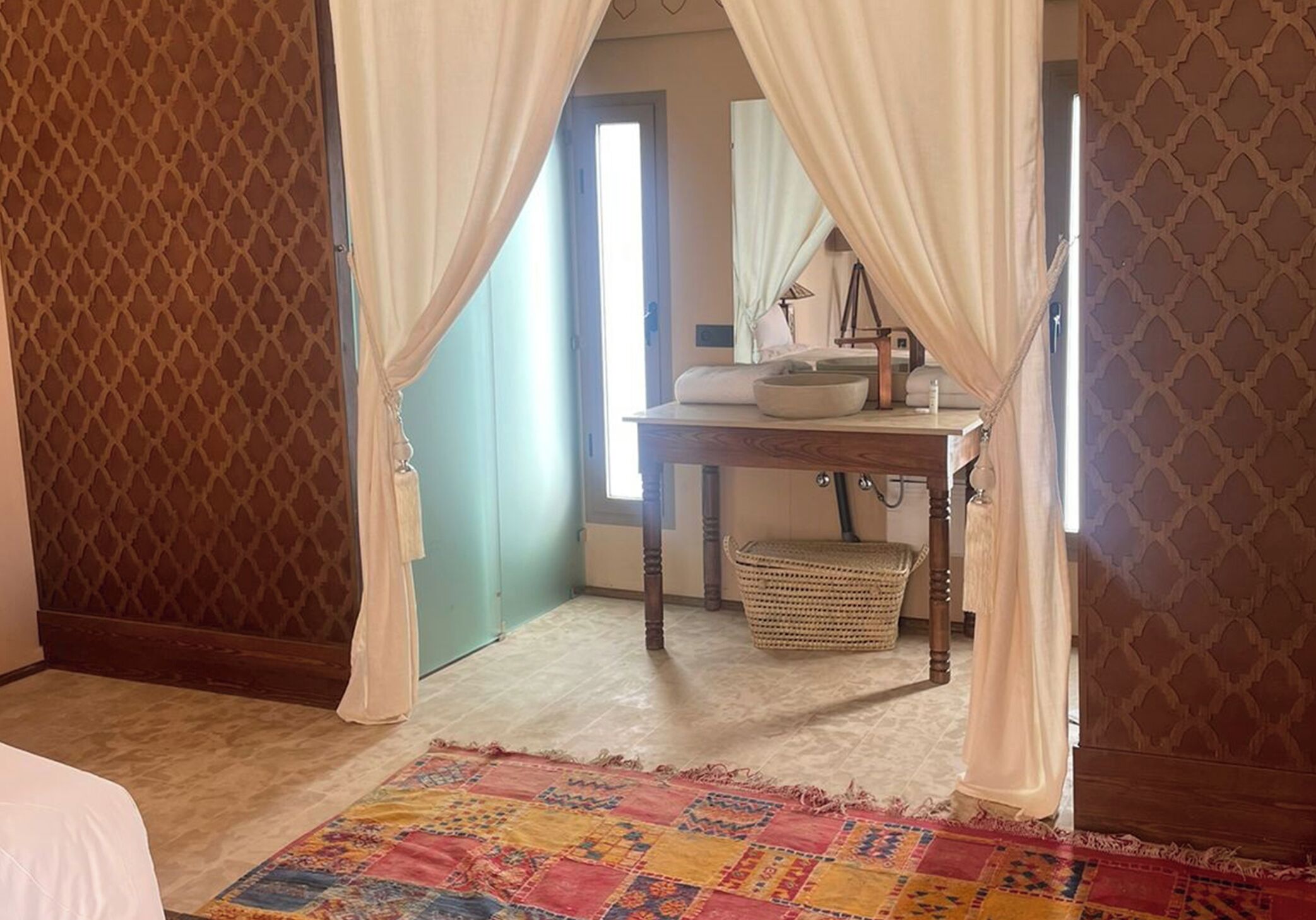 A room with a bed, a bedside table with a lamp, a window with curtains, and a patterned rug.