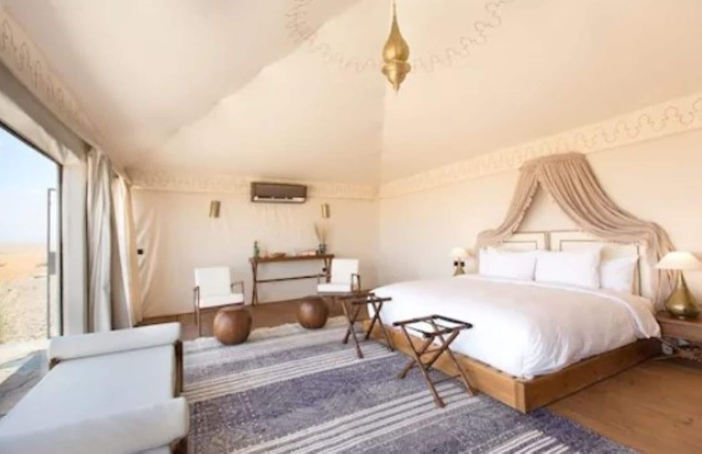 A bedroom with a large bed, a desk, and a chair. There is a window with curtains, a hanging light fixture, and a patterned rug.