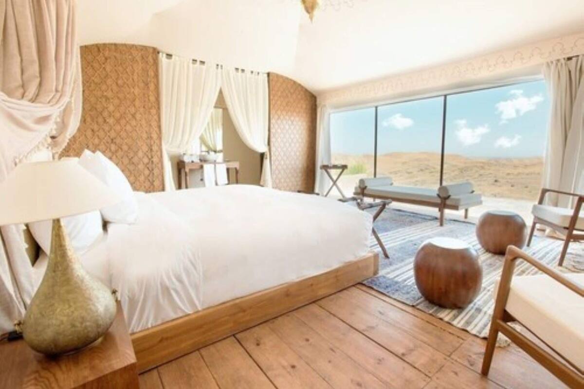 A bedroom with a large bed, a bedside table, a lamp, a window with a view of the desert, and a seating area with a bench and ottoman.