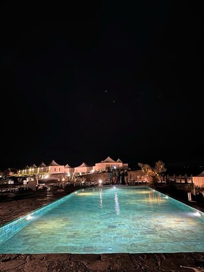 A nighttime view of a pool area with a clear pool, a series of lighted buildings in the background, and a few people standing by the pool. - The White Camel Lodge (Agafay)