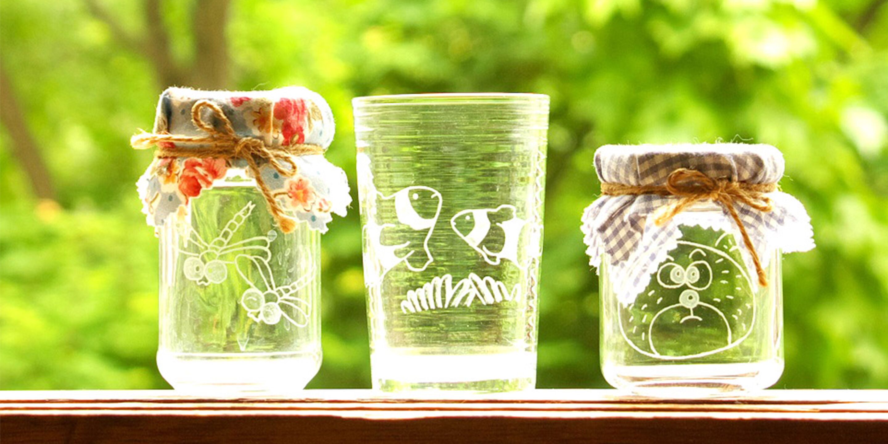Three glass jars with different designs, tied with fabric and twine, placed on a wooden surface with a blurred green background.