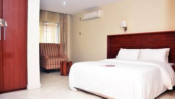 A hotel room with a large bed, a wooden headboard, an air conditioning unit, a striped armchair, and a small wooden table.
