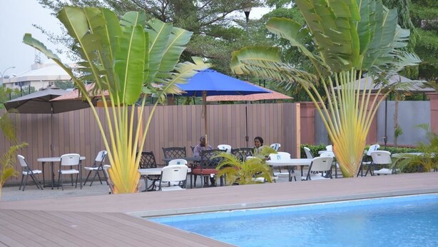 Un espace extérieur avec une piscine, une terrasse en bois, des chaises et des tables blanches. De grands bananiers et des parasols offrent de l’ombre.
