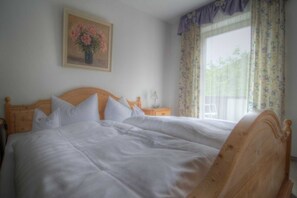 Ein Doppelbett mit weißer Bettwäsche, ein Holzkopfschild, ein Nachttisch mit Lampe und ein Fenster mit blumigen Vorhängen.