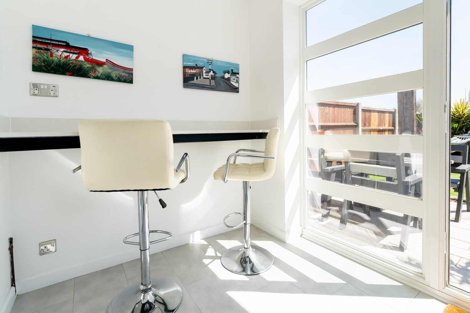 A room with two bar stools, a large window, and two paintings on the wall.