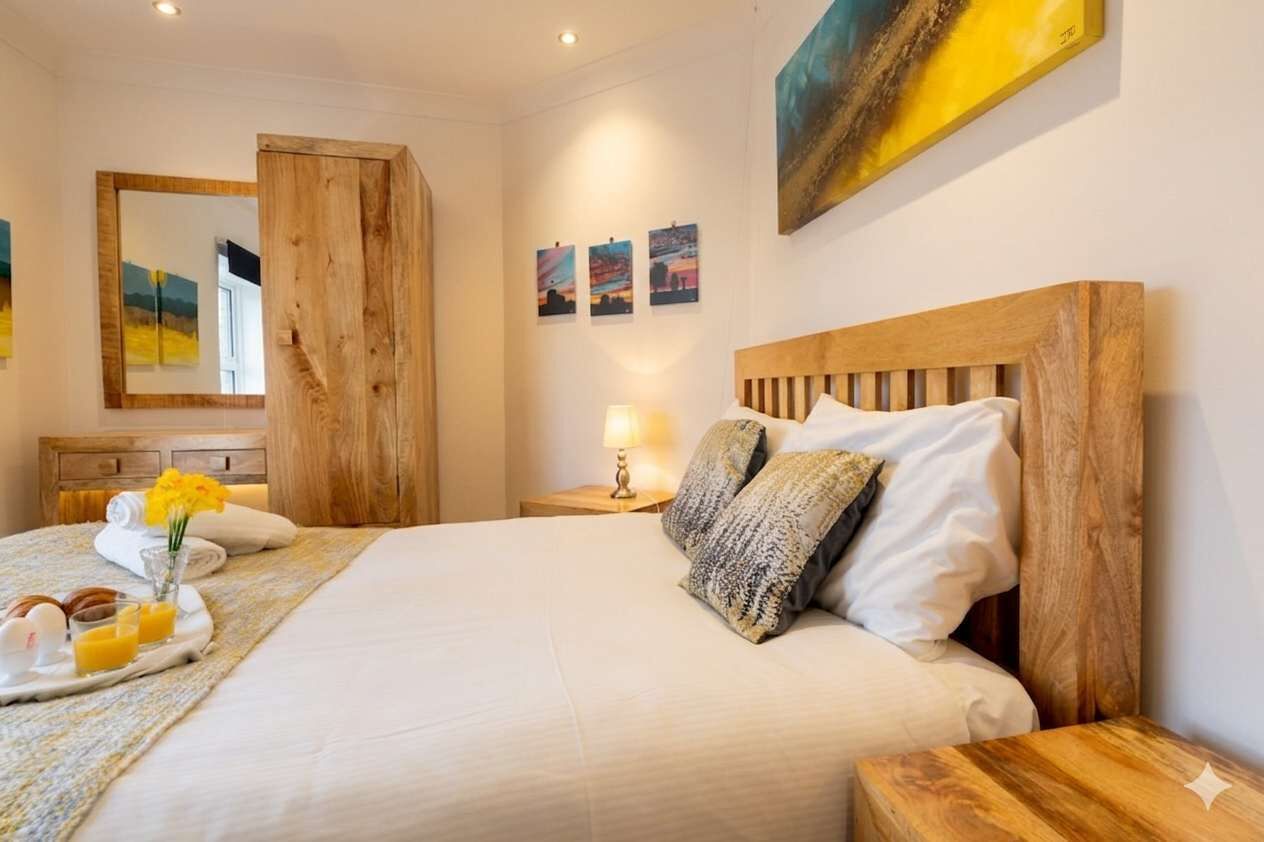 A hotel room with a large bed, wooden headboard, a bedside table with a lamp, a mirror, and artwork on the wall.