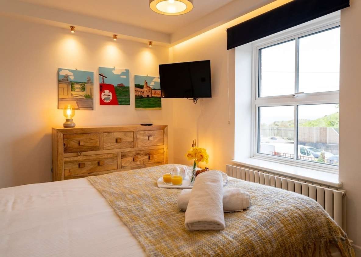 A hotel room with a bed, wooden dresser, artwork on the wall, a TV, and a window with a view.