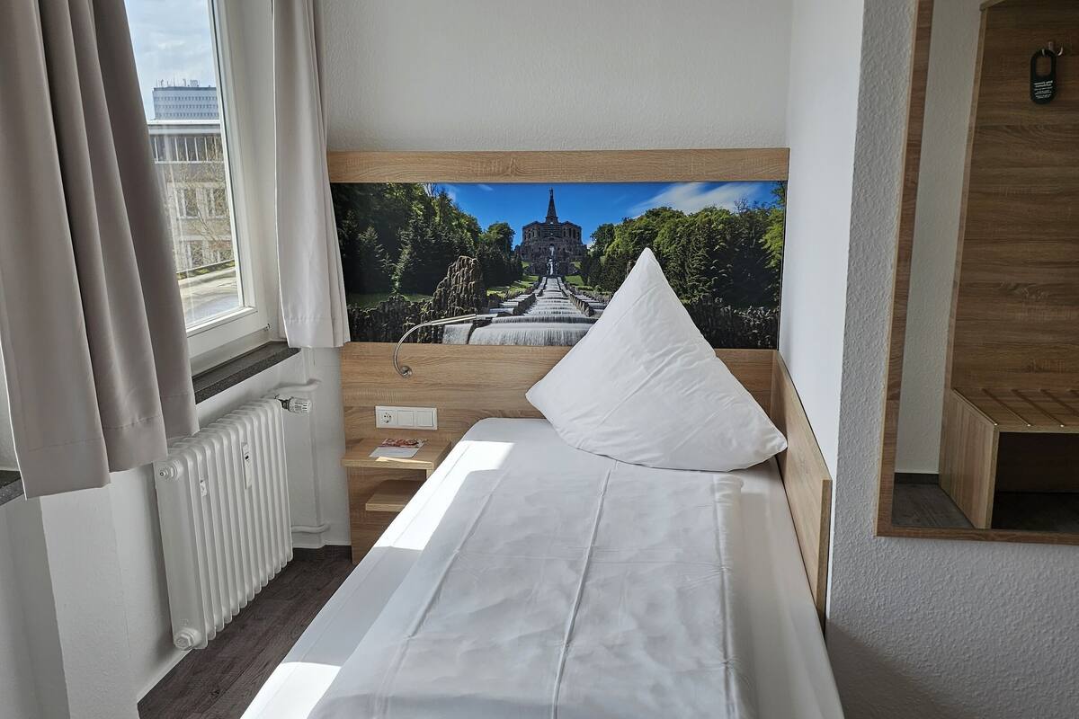 A hotel room with a bed, a window with curtains, a radiator, a nightstand, and a wall-mounted TV.