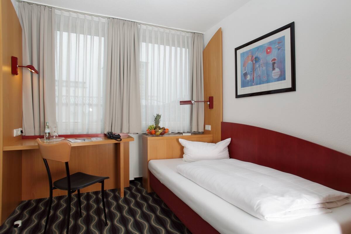 A hotel room with a bed, a desk, a chair, a window with curtains, a wall-mounted artwork, and a small table with fruits.