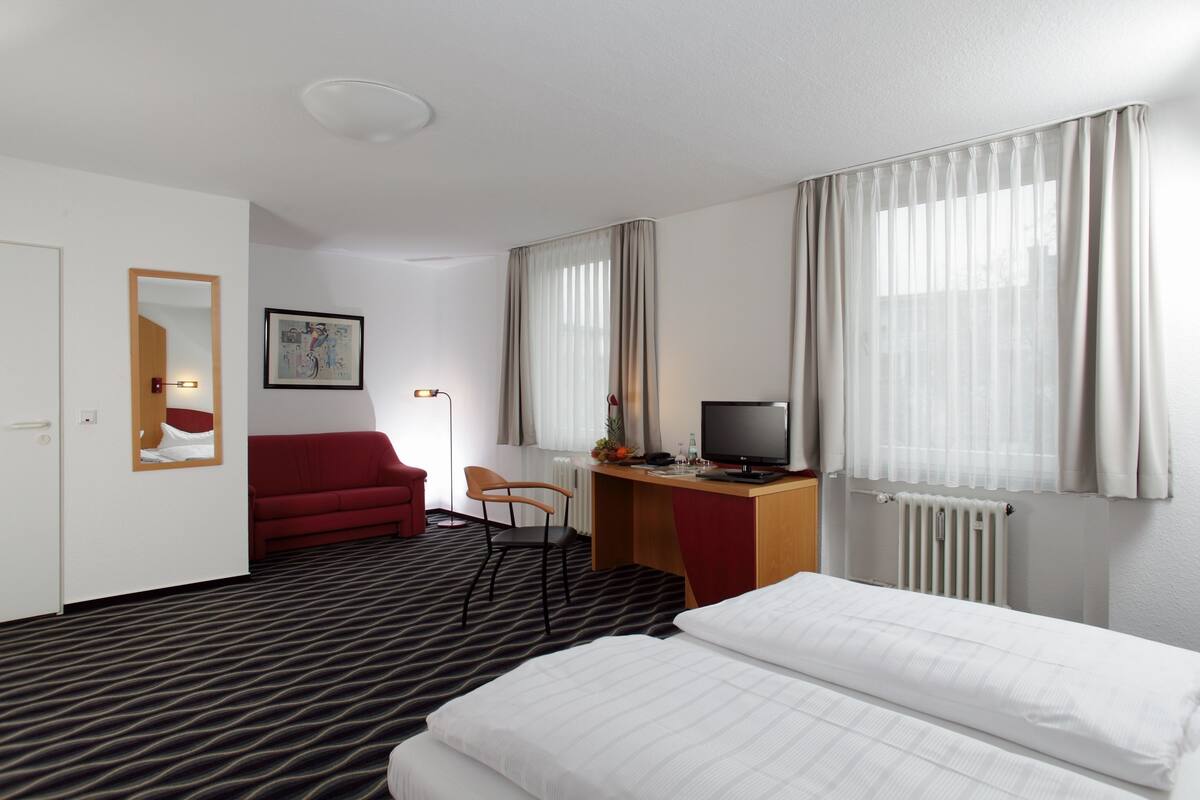 A hotel room with a bed, a desk with a computer, a red sofa, a chair, a mirror, and a framed picture on the wall.