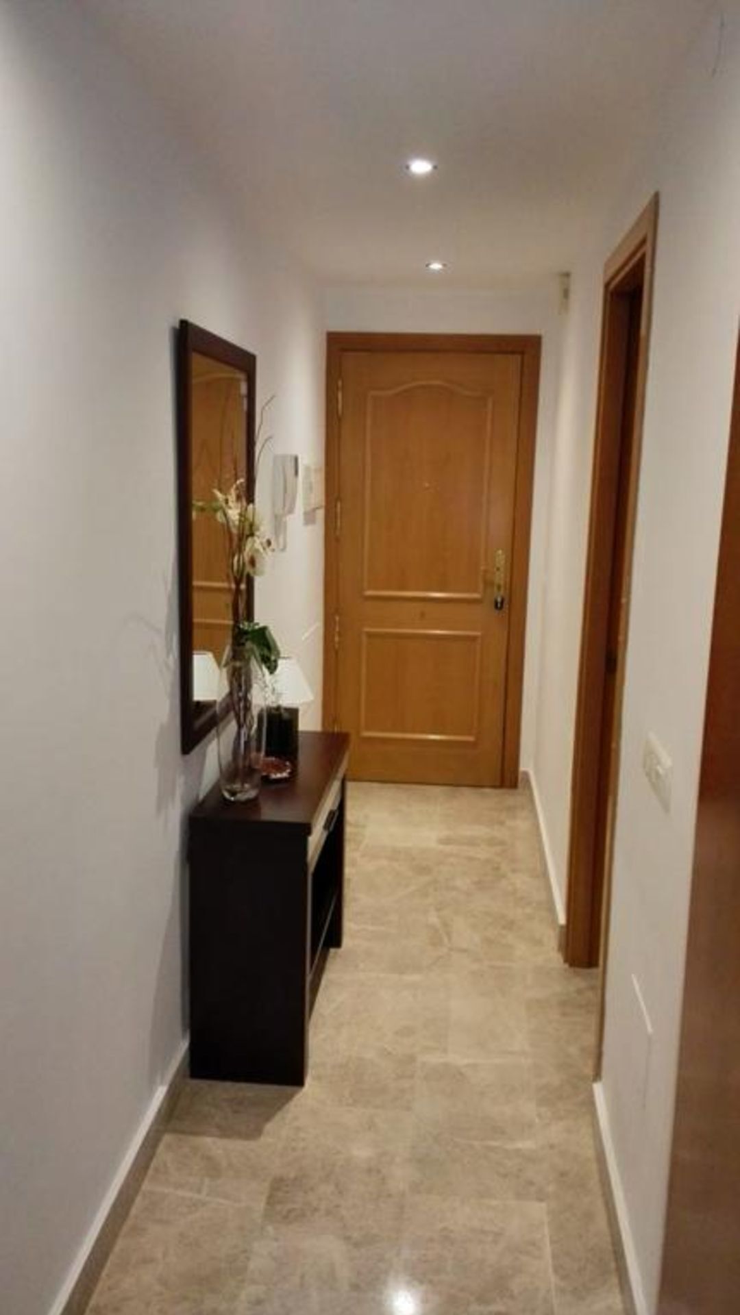 A hallway with a wooden door, a dark console table with a mirror and a plant, and a tiled floor.