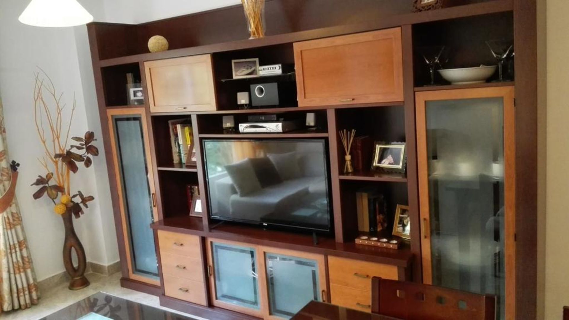 A living room with a flat-screen TV, wooden cabinets, and a glass-fronted cabinet.