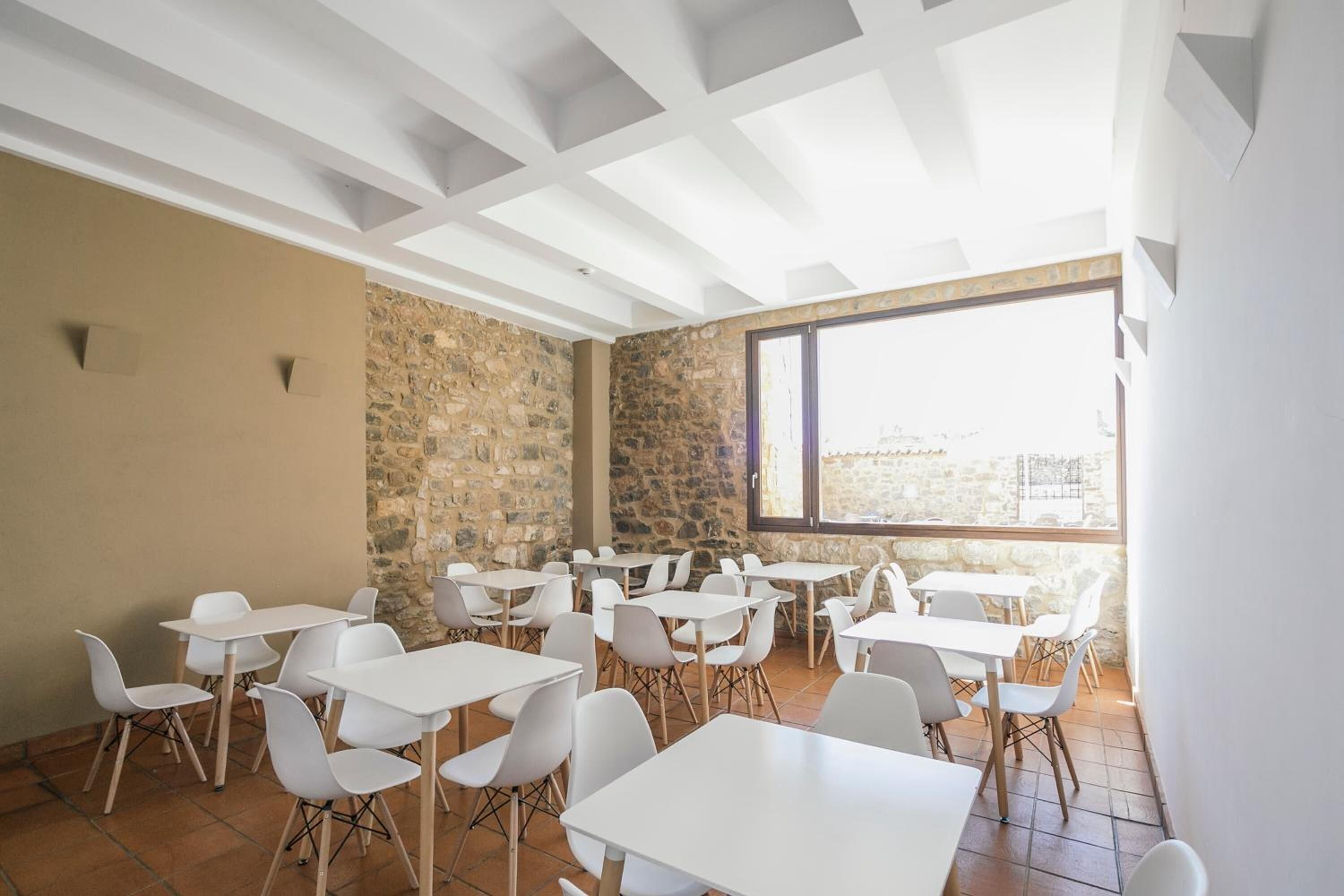 Une pièce spacieuse avec des tables et des chaises blanches, un mur en pierre et une grande fenêtre donnant sur un beau paysage.