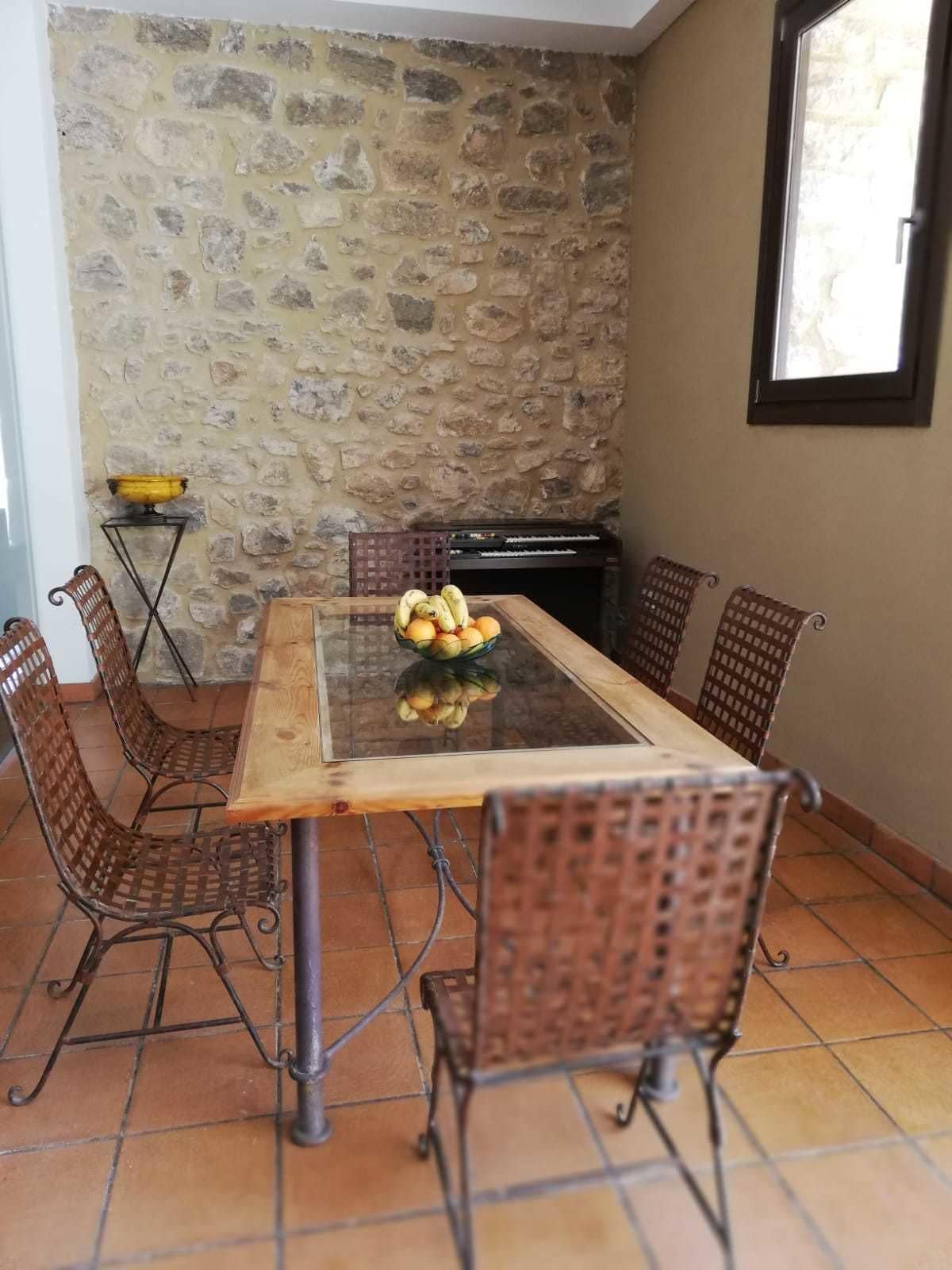 Un coin repas agrémenté d’un mur en pierre, d’une table à plateau en verre, de chaises en osier et d’une petite table sur laquelle est posé un bol.
