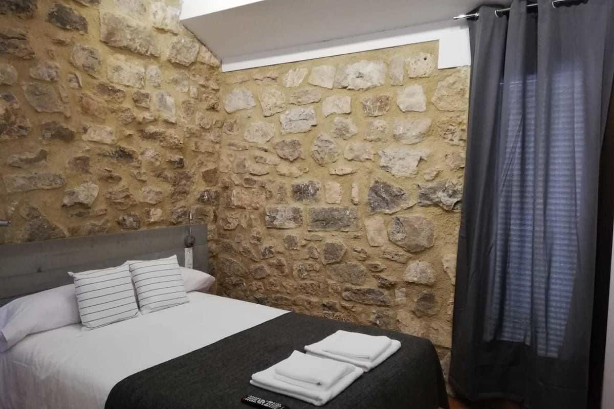 A bedroom with a stone wall, a bed with white bedding, and a remote control on the bed.