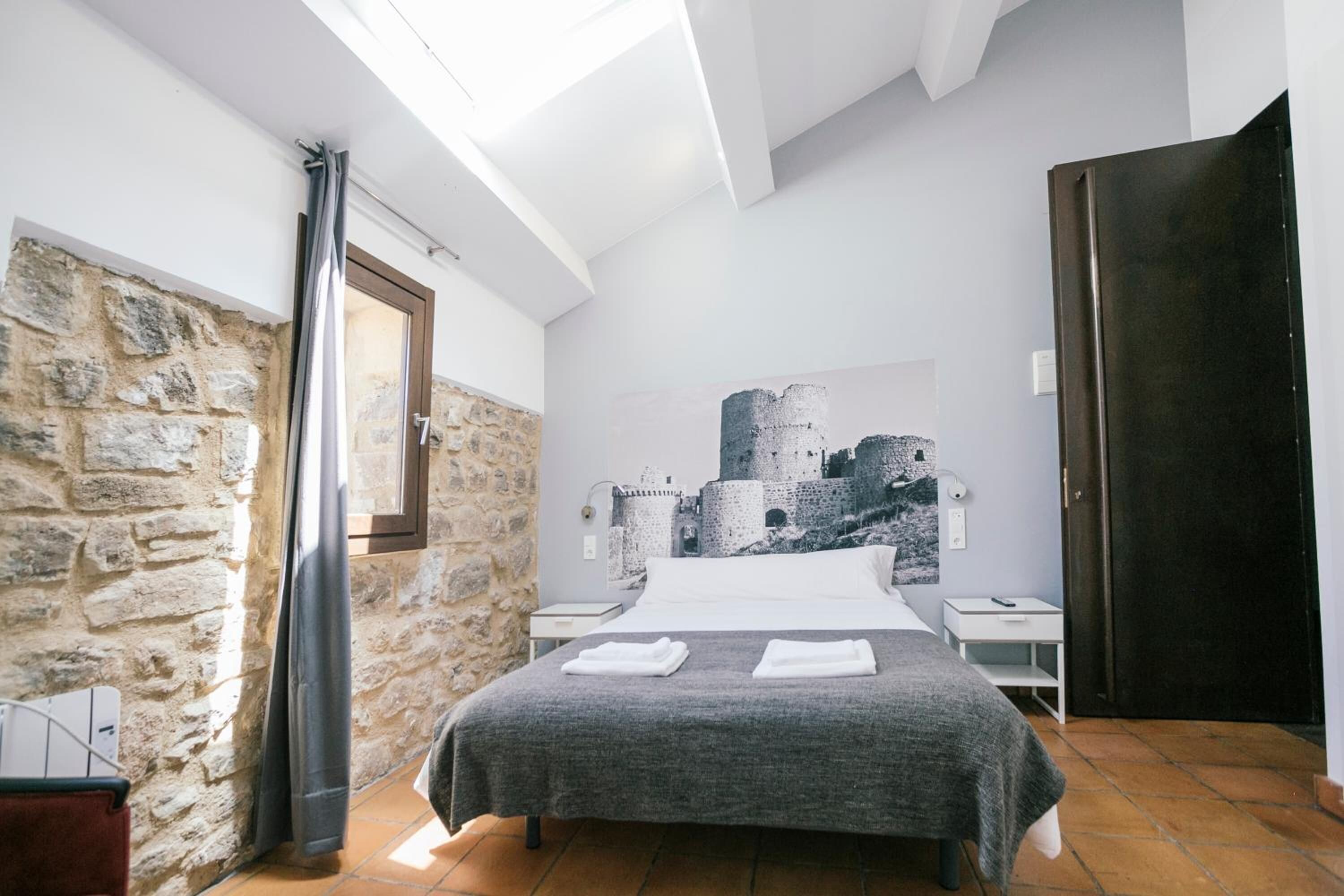 Een slaapkamer met een stenen muur, een bed met een grijze deken, een nachtkastje, een houten deur en een muurschildering van een kasteel op de muur.